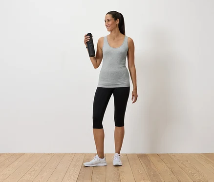 Frau hält eine Wasserflasche, trägt ein graues Tanktop, schwarze Leggings und weiße Turnschuhe.