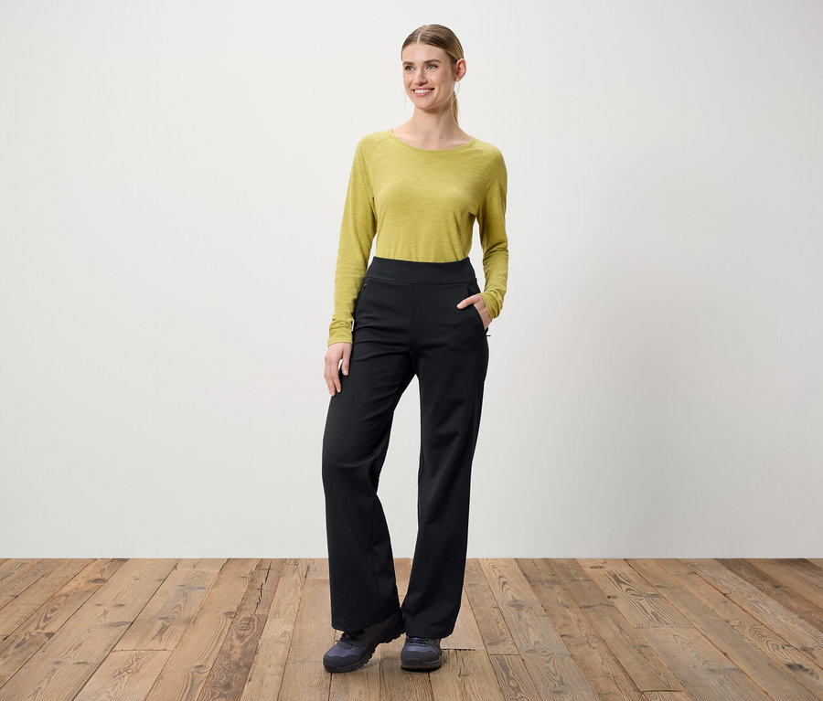 Eine Frau steht auf einem Holzboden. Sie trägt eine grüne Bluse mit langen Ärmeln und eine schwarze Hose.