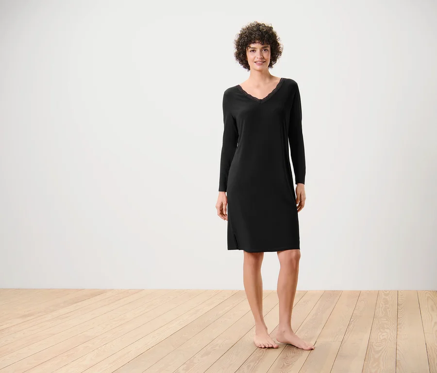 Eine Frau steht barfuß auf einem Holzboden und trägt ein schwarzes, langärmliges Kleid.