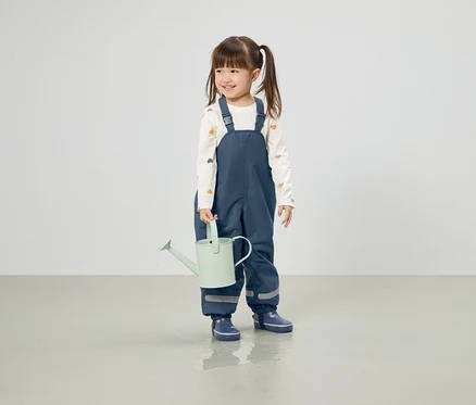 Lächelndes Mädchen mit Zöpfen hält eine hellgrüne Gießkanne, gekleidet in blaue Hosen und Gummistiefel.