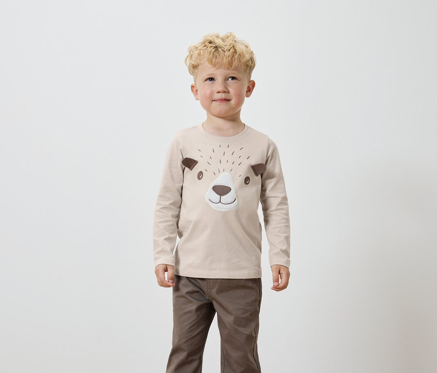 Ein Junge mit blond gelocktem Haar trägt ein beiges Langarmshirt mit braunen Bärengesicht-Applikationen und braune Hosen.