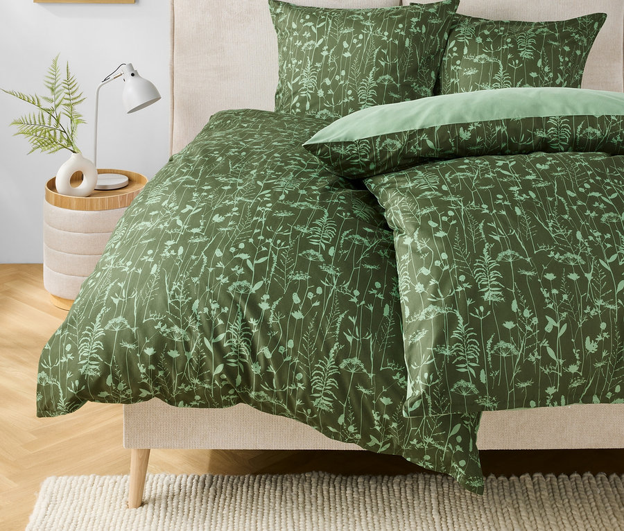 Grüne Bettwäsche mit Blumenmuster auf einem Bett. Auf dem Nachttisch stehen eine Lampe und eine Vase mit einer Pflanze.