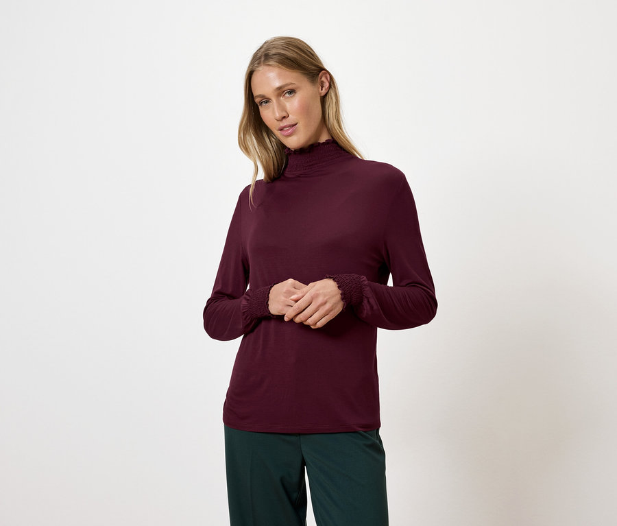 Blonde Frau trägt ein weinrotes Rollkragenshirt und grüne Hosen.