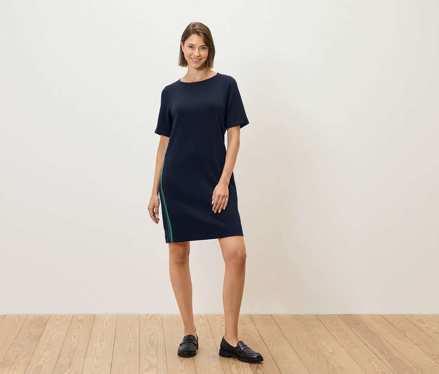Eine Frau steht und trägt ein dunkelblaues Kleid mit kurzen Ärmeln und einem grünen Streifen an der Seite.