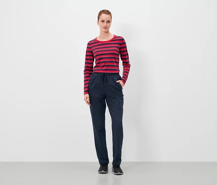 Frau steht mit Händen in den Taschen einer dunkelblauen Hose und trägt ein rot-blau gestreiftes Langarmshirt.