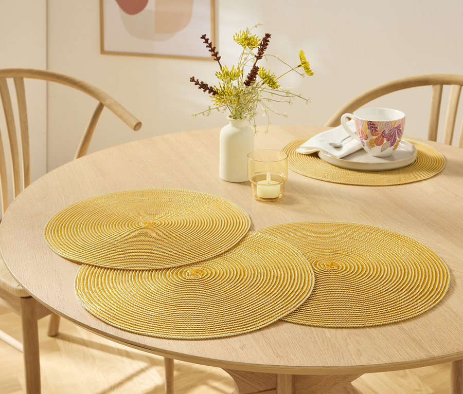 Auf einem hellen Holztisch liegen drei gelbe, runde Platzsets, eine Vase mit Blumen, eine Kerze und eine Tasse mit Untertasse.