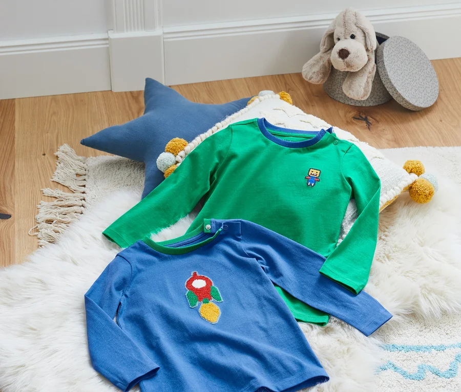 Zwei Langarmshirts liegen auf einem weißen zotteligen Teppich neben einem blauen Stern und einem Spielzeug.