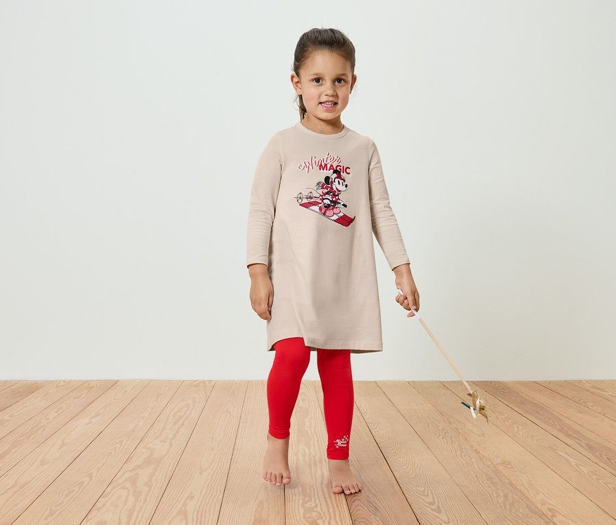 Mädchen in einem beigen Kleid mit Minnie-Mouse-Motiv und roten Leggings steht auf einem Holzboden.