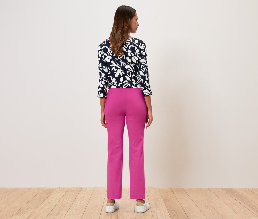 Frau steht mit dem Rücken zur Kamera, trägt eine geblümte Bluse und pinke Hosen.