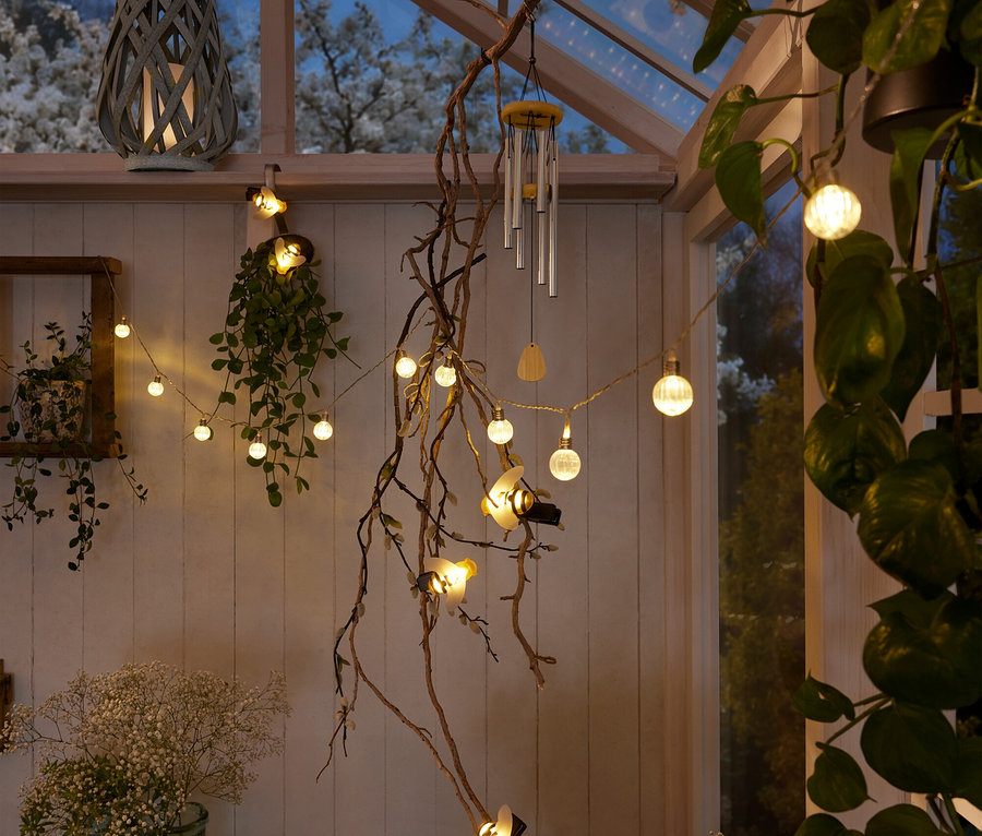 Innenaufnahme eines Wintergartens mit einer grauen Laterne, einer Lichterkette mit Kugeln und Bienen, einem Windspiel und Pflanzen.