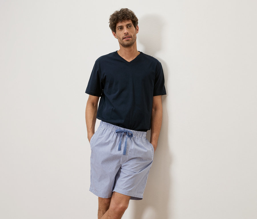 Mann mit lockigem Haar trägt ein marineblaues T-Shirt und gestreifte Shorts, während er an einer weißen Wand lehnt.