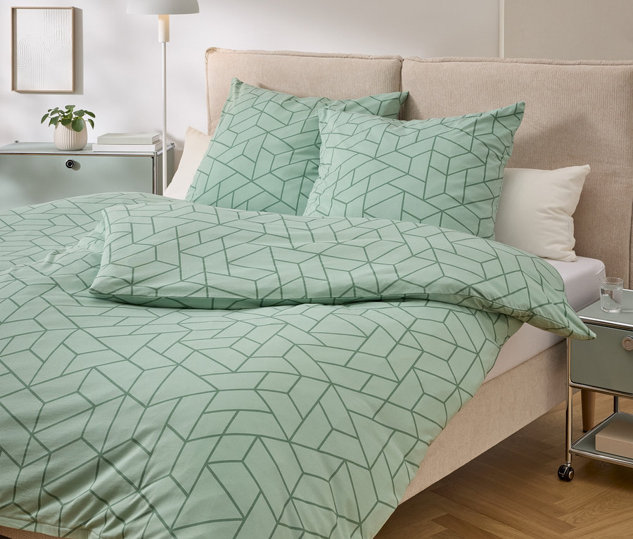 Bett mit mintgrüner Bettwäsche mit geometrischem Muster, zwei Kissen, Nachttisch und Lampe.