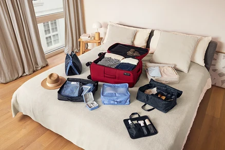 Ein roter Koffer und diverse Packtaschen liegen auf einem Bett mit einem Überwurf aus gewaschenem Leinen.
