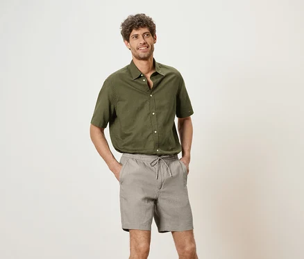 Mann mit olivgrünem Hemd und grauen Shorts, die Hände in den Hosentaschen, vor einem neutralen Hintergrund.