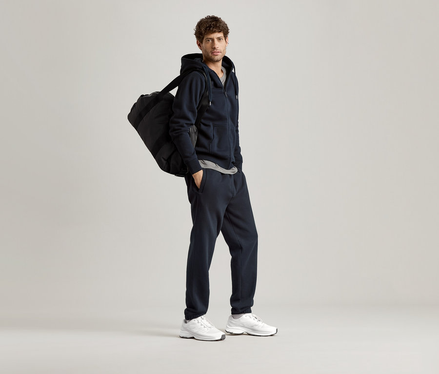 Mann mit marineblauer Kapuzenjacke und Sporttasche über der Schulter.