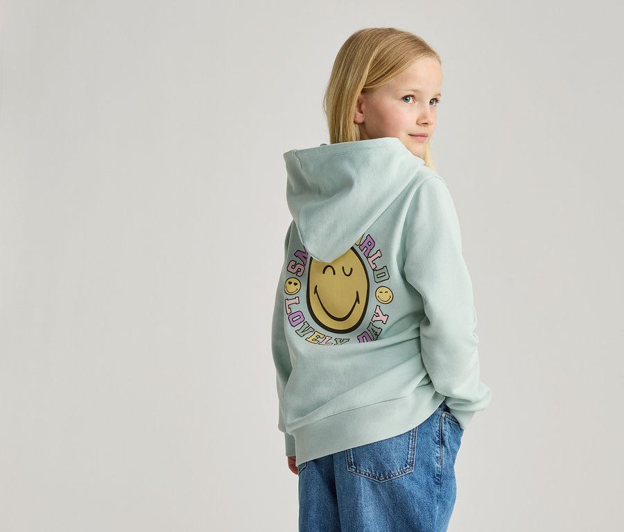 Mädchen mit blonden Haaren, von hinten gesehen, trägt einen hellblauen Kapuzenpullover mit Smiley-Aufdruck und Jeans.