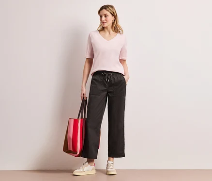 Frau mit rosa Oberteil und Street One Papertouch-Hose posiert mit einer rot gestreiften Tasche.