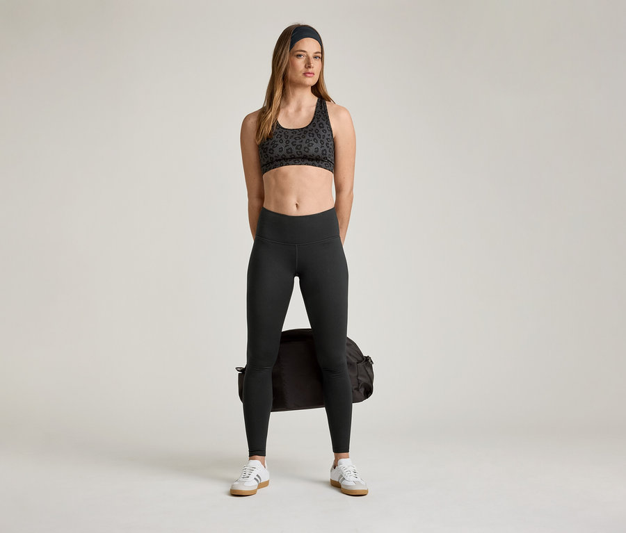 Frau in Sport-BH und »ActiveSoft« Sportleggings mit Sporttasche und Turnschuhen.