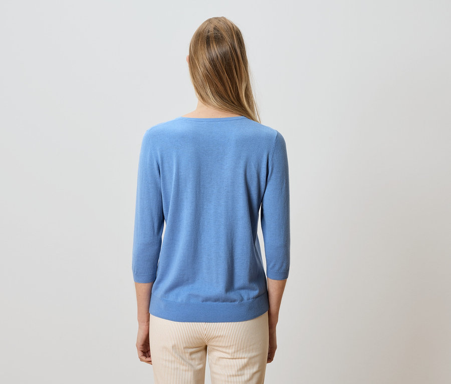 Rückenansicht einer Frau mit blonden, langen Haaren, die einen hellblauen Pullover und gestreifte Hosen trägt.