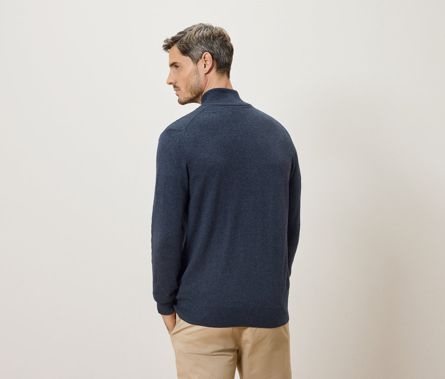 Mann von hinten in marineblauem Pullover und beigen Hosen vor einem cremefarbenen Hintergrund.