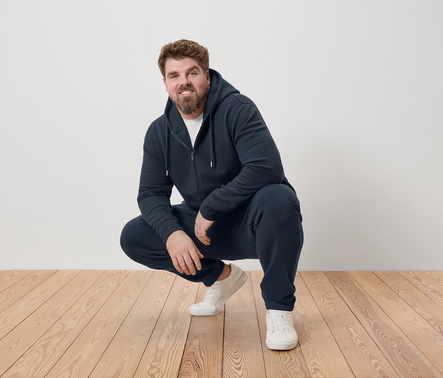 Mann hockt auf einem Holzboden, trägt einen dunkelblauen Trainingsanzug und weiße Turnschuhe.