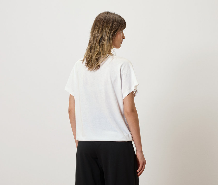 Eine Frau mit gewelltem, schulterlangem Haar trägt ein weißes T-Shirt mit Spitzenapplikationen und eine schwarze Hose.