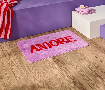Auf einem Holzboden liegt eine lila Badematte mit dem roten Schriftzug "AMORE".
