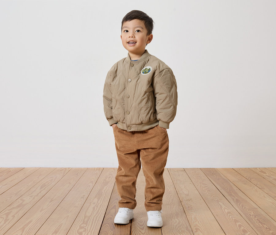 Ein lächelnder Junge mit beiger, gesteppter Jacke mit Aufnäher, Cordhose und weißen Schuhen steht auf einem Holzboden vor einer weißen Wand.