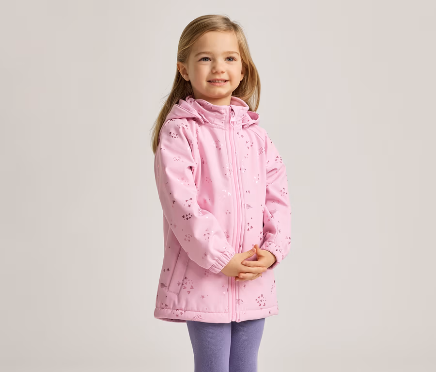Ein junges Mädchen mit blonden Haaren lächelt und trägt eine rosa, gemusterte Jacke und lila Leggings.