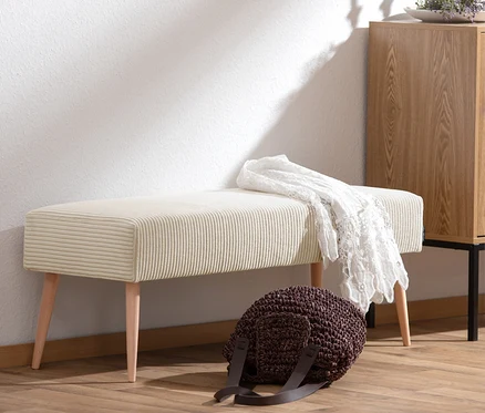 Eine cremefarbene Sitzbank »Sit« aus Cordstoff steht vor einer weißen Wand. Eine weiße Häkeldecke liegt auf der Bank. Eine braune Tasche steht davor.