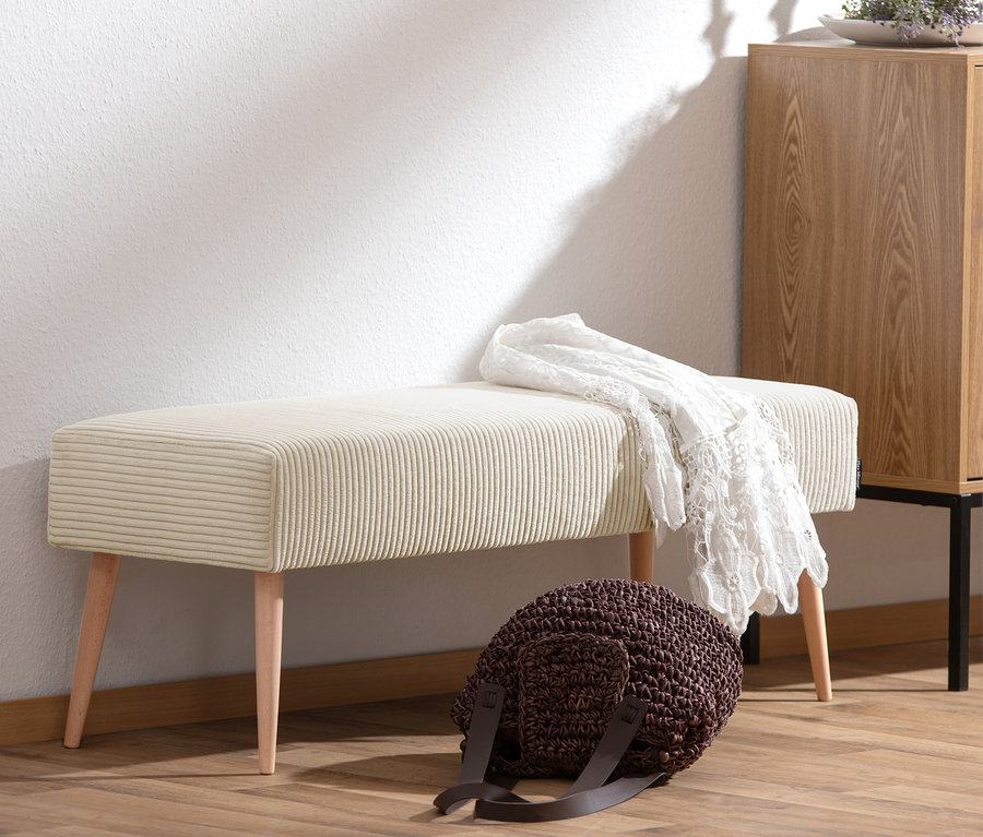 Eine cremefarbene Sitzbank »Sit« aus Cordstoff steht vor einer weißen Wand. Eine weiße Häkeldecke liegt auf der Bank. Eine braune Tasche steht davor.