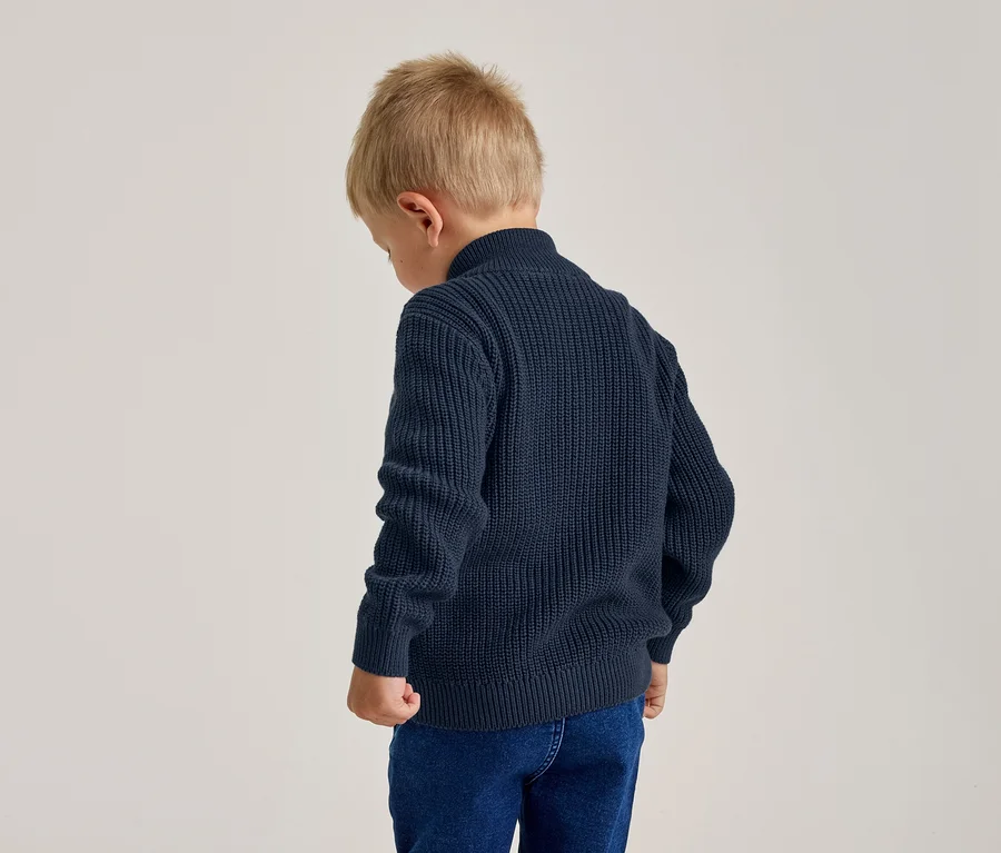Junger Junge von hinten, trägt einen marineblauen Strickpullover und blaue Jeans, der Kopf ist leicht gesenkt.