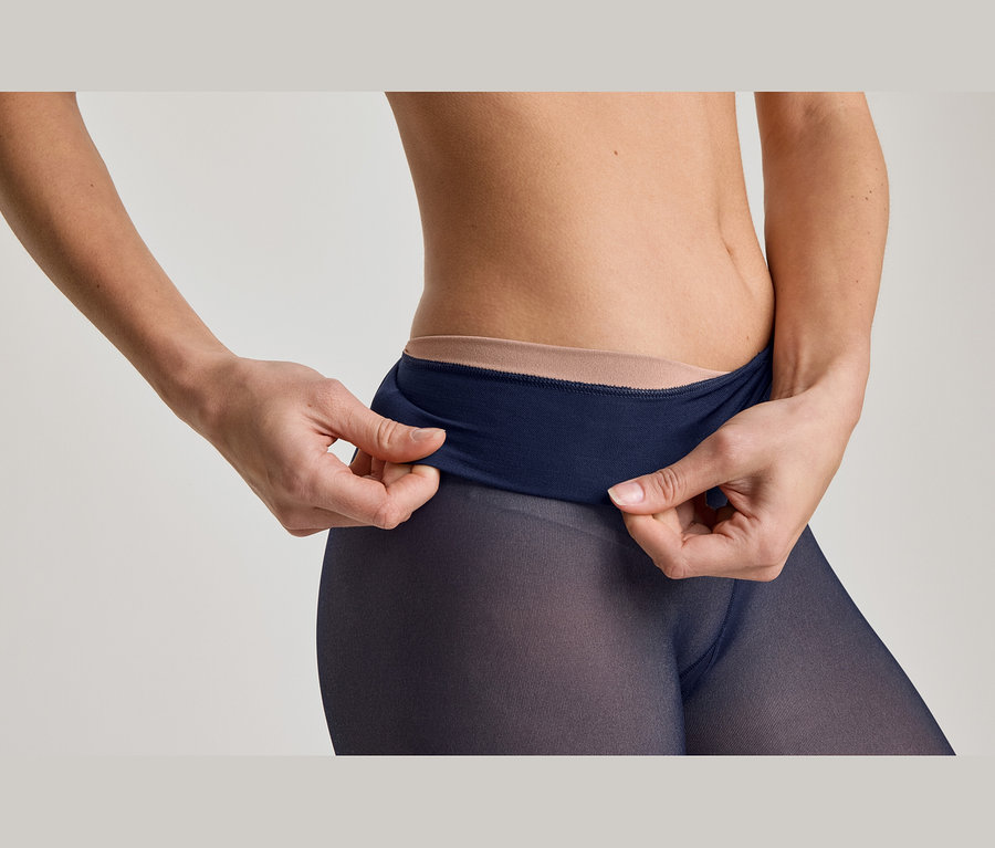 Nahaufnahme einer Person, die den breiten, dunkelblauen Bund einer transparenten, dunkelblauen Strumpfhose hochzieht. Darunter ist ein hellerer, hautfarbener Bund sichtbar.