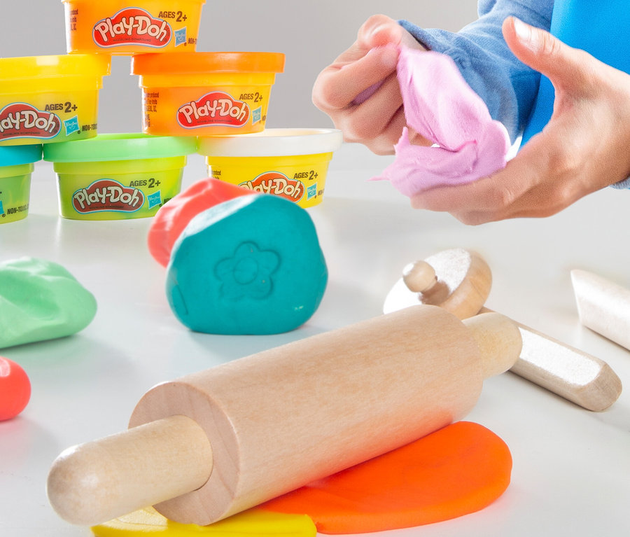 Hände formen rosa Play-Doh. Im Vordergrund liegen bunte Play-Doh-Klumpen, ein Nudelholz und Play-Doh-Dosen im Hintergrund.