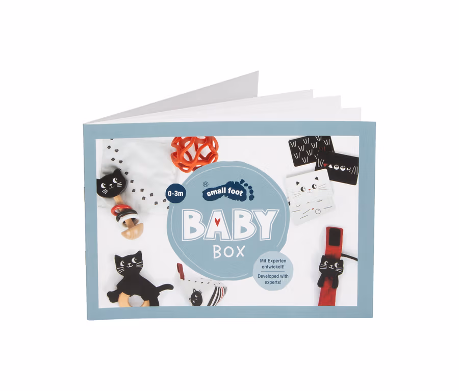 Eine offene small foot Baby-Box, 0–3 Monate, gefüllt mit verschiedenen Babyartikeln wie einer schwarzen Katzenrassel, einem roten Greifling, schwarz-weißen Stoffkarten und einem Beißring.