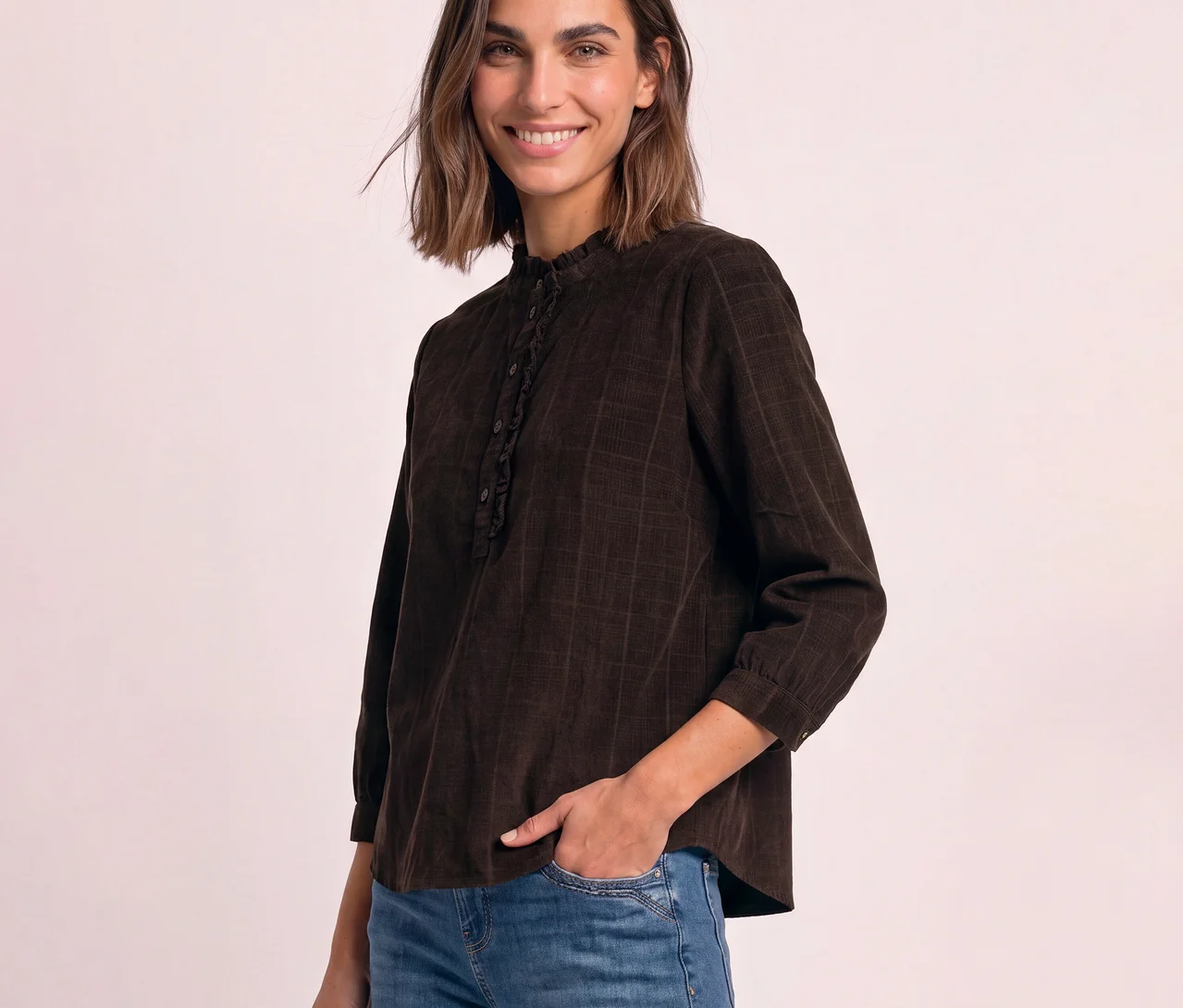 Lächelnde Frau trägt eine braune Street One Cordbluse mit Rüschen-Stehkragen und Knopfleiste sowie eine blaue Jeans, die linke Hand steckt in der Hosentasche.
