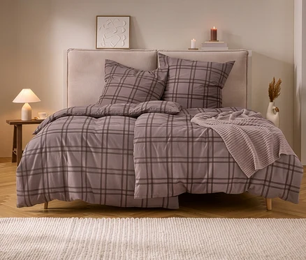 Doppelbett mit karierter Biber-Bettwäsche in Grau-Braun und beigem Stricküberwurf. Daneben ein Nachttisch mit Lampe und Kerzen.