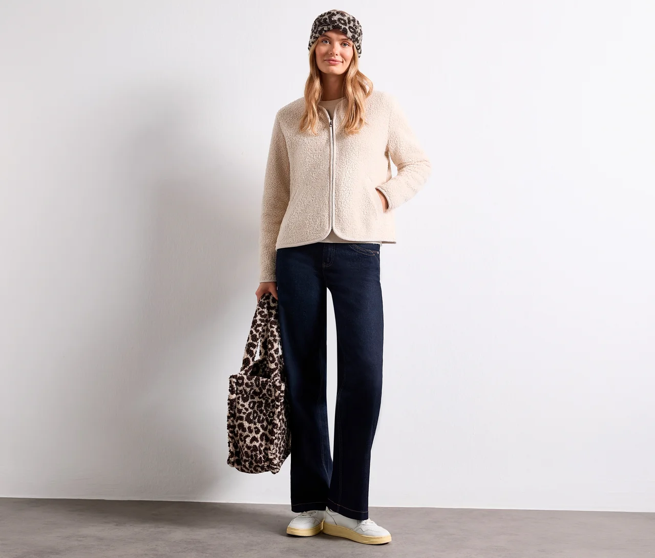 Eine Frau trägt ein beiges Teddyfell-Jäckchen, eine dunkle Jeans, weiße Sneaker und ein Street One Stirnband mit Leopardenmuster. Sie hält eine passende Tasche.