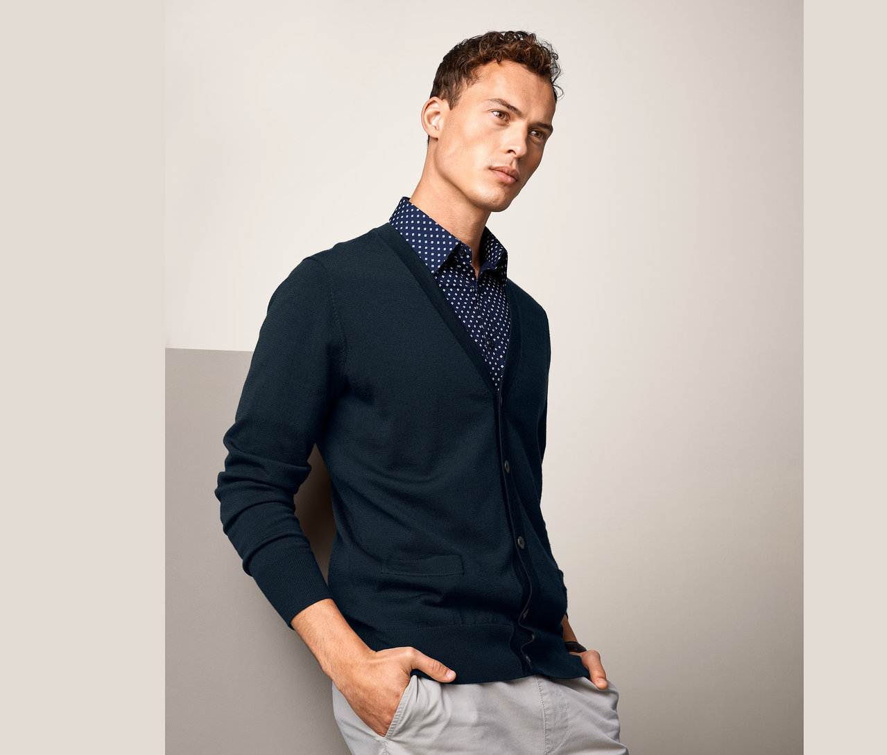 Mann lehnt an Wand, trägt marineblauen Merino-Cardigan und blaues Hemd mit Punkten.