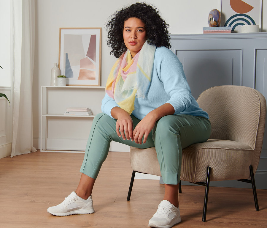 Frau sitzt auf einem Stuhl und trägt einen hellblauen Strickpullover, eine mintfarbene Stretchhose, einen Webschal und Veloursleder-Sneaker.