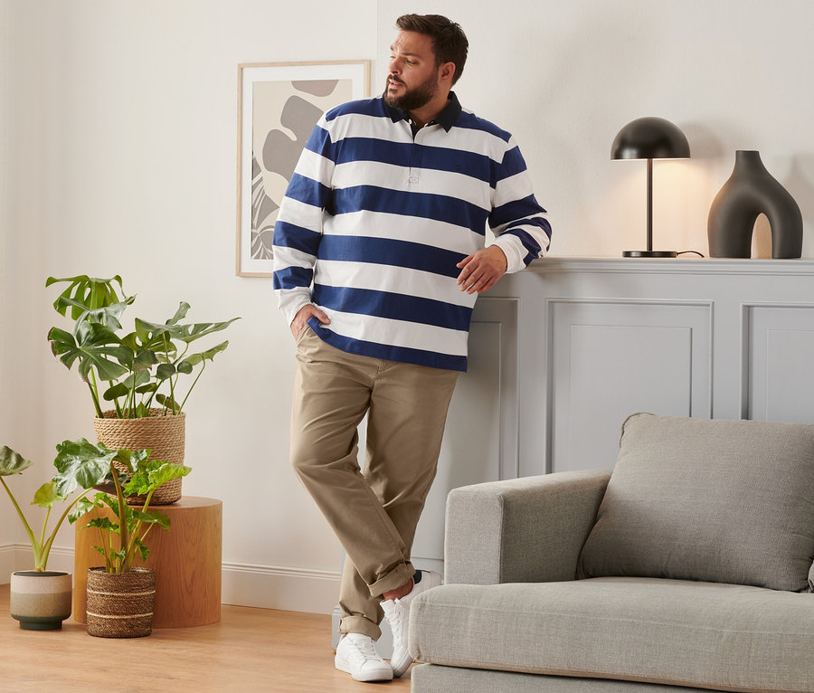 Mann lehnt an Wand, trägt camelfarbene Chino, blau-weißes Langarm-Poloshirt und Sneaker.