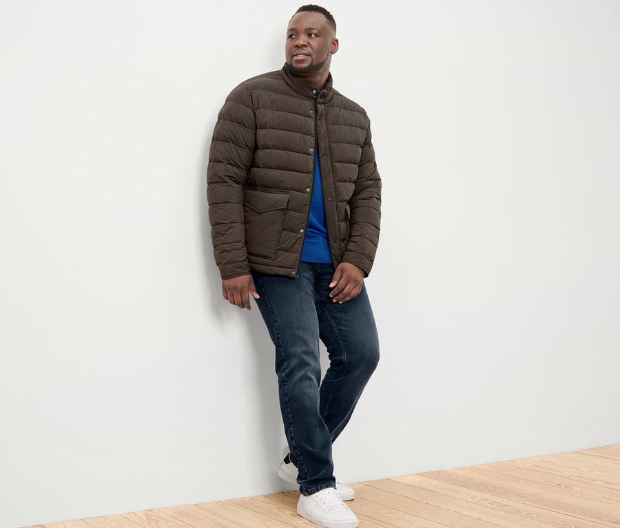 Mann an eine Wand gelehnt trägt eine Steppjacke, Jeans und Turnschuhe.