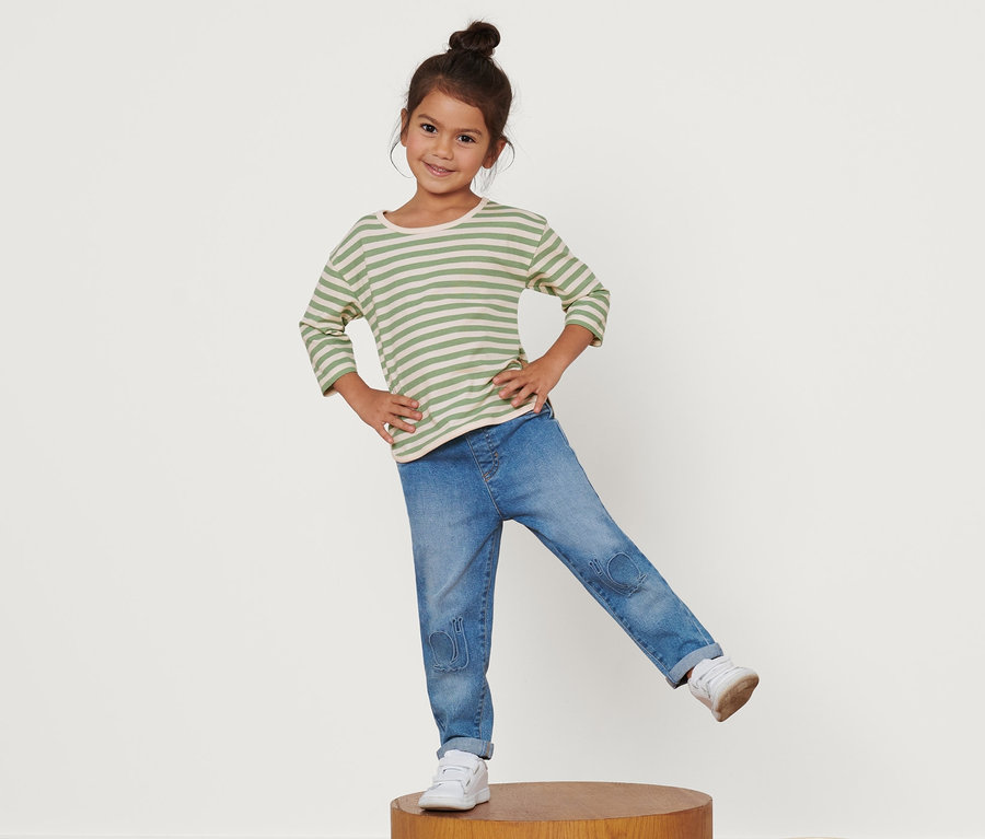 Ein Mädchen steht auf einem Holzsockel und trägt ein Kinder-Langarmshirt, Jeans und Turnschuhe.