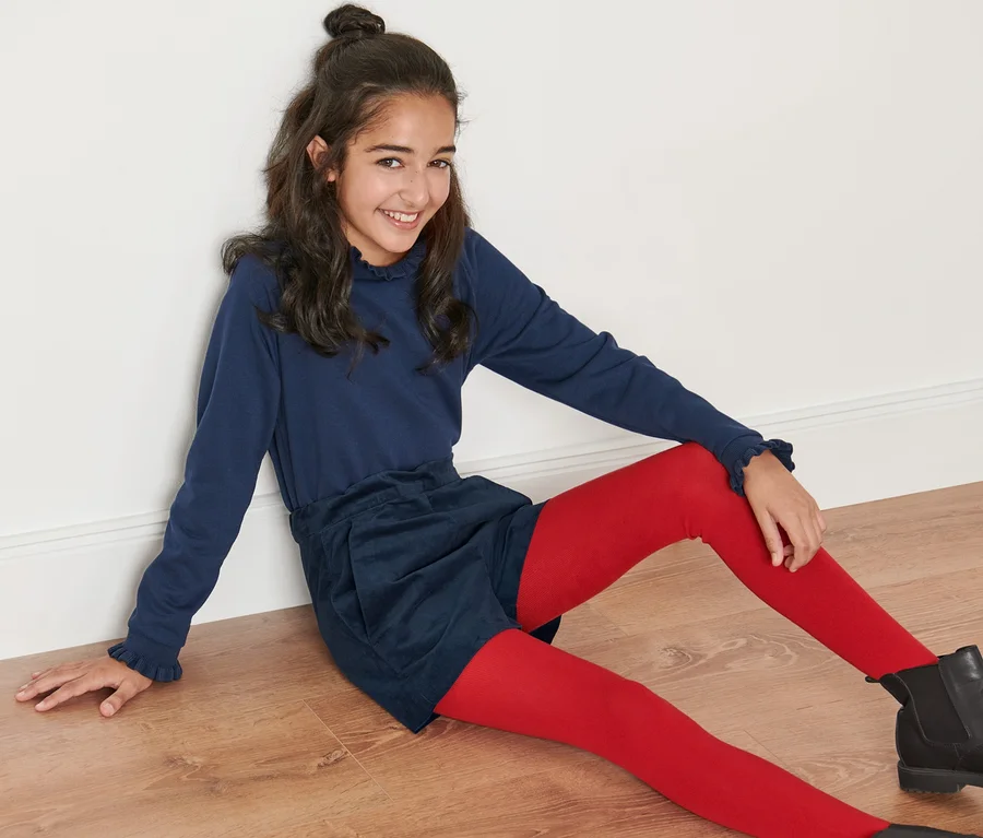 Ein lächelndes Mädchen sitzt auf dem Boden und trägt Kinder-Samtshorts, eine rote Strumpfhose und schwarze Stiefel.