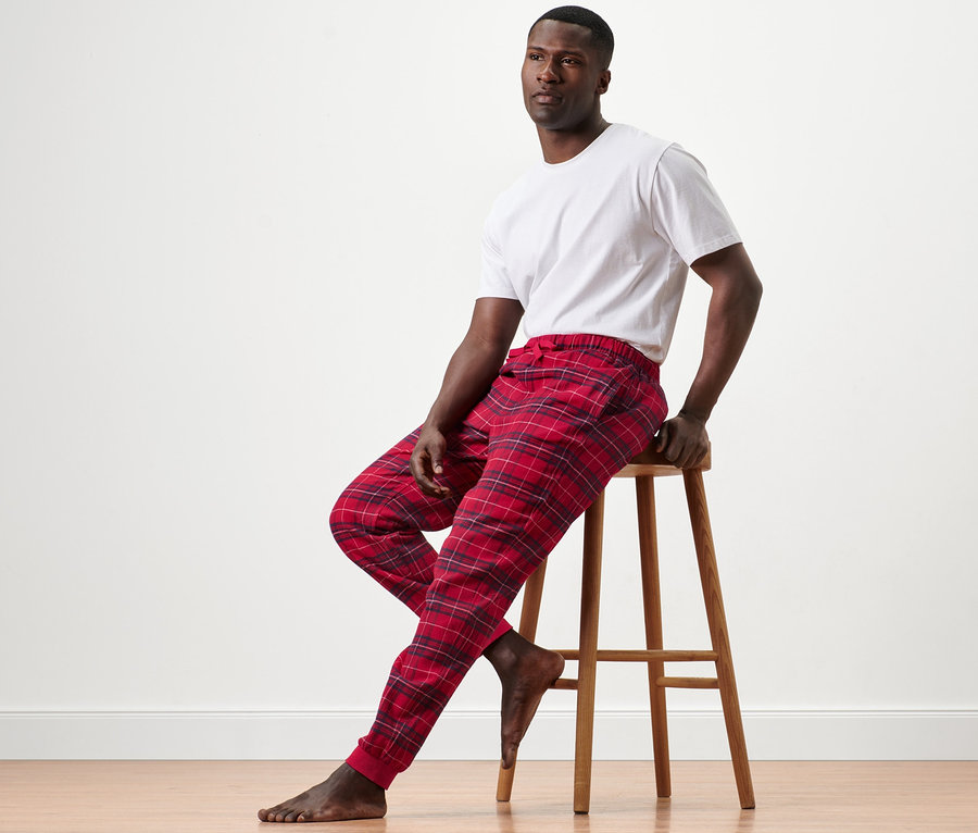 Mann sitzt auf einem Holzhocker und trägt eine Flanell-Relaxhose und ein weißes T-Shirt.