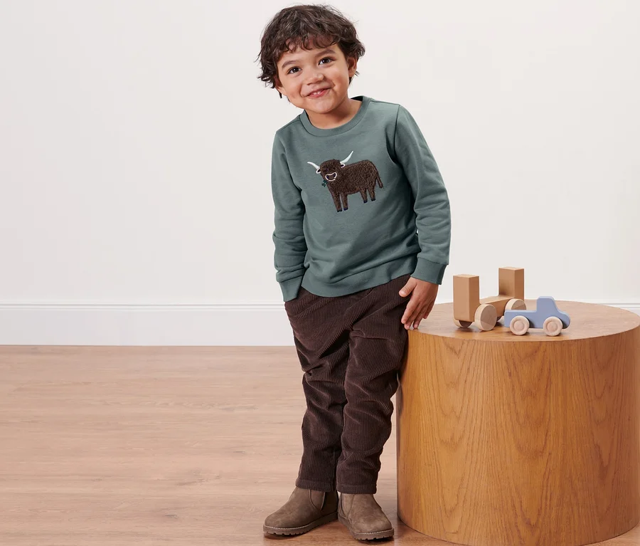 Junge in Kleinkind-Sweater mit Hochlandrind-Motiv lehnt an rundem Tisch mit Holzspielzeug.