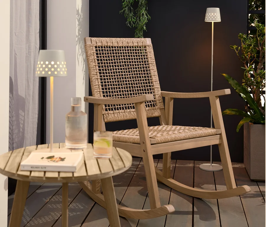 Auf einer Terrasse stehen ein Schaukelsessel mit Polyrattangeflecht, ein Beistelltisch »Lenja« mit 2 Ebenen in Teakoptik, eine LED-Akku-Tischleuchte aus Metall und eine LED-Akku-Stehleuchte aus Metall.