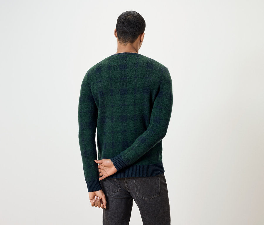 Ein Mann mit dem Rücken zur Kamera trägt einen grün karierten Pullover und dunkle Jeans.