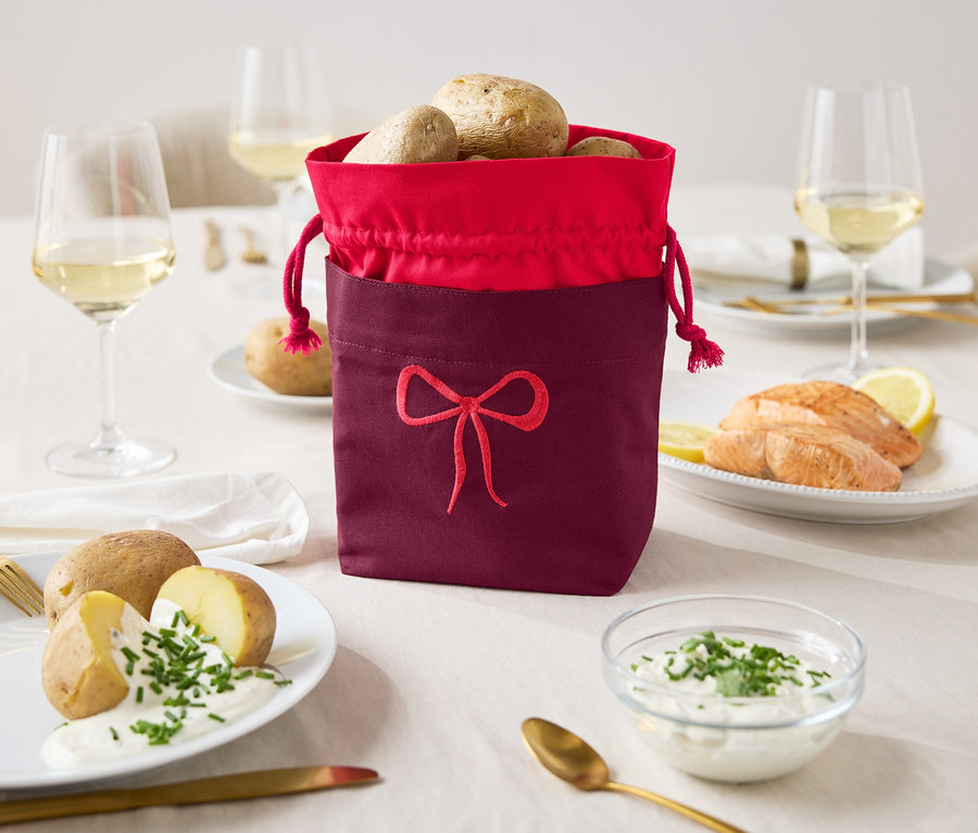 Auf einem Tisch stehen ein weinroter Stoffbeutel mit rotem Rand, gefüllt mit Kartoffeln, neben Tellern mit Kartoffeln mit Soße und Lachs mit Zitrone.