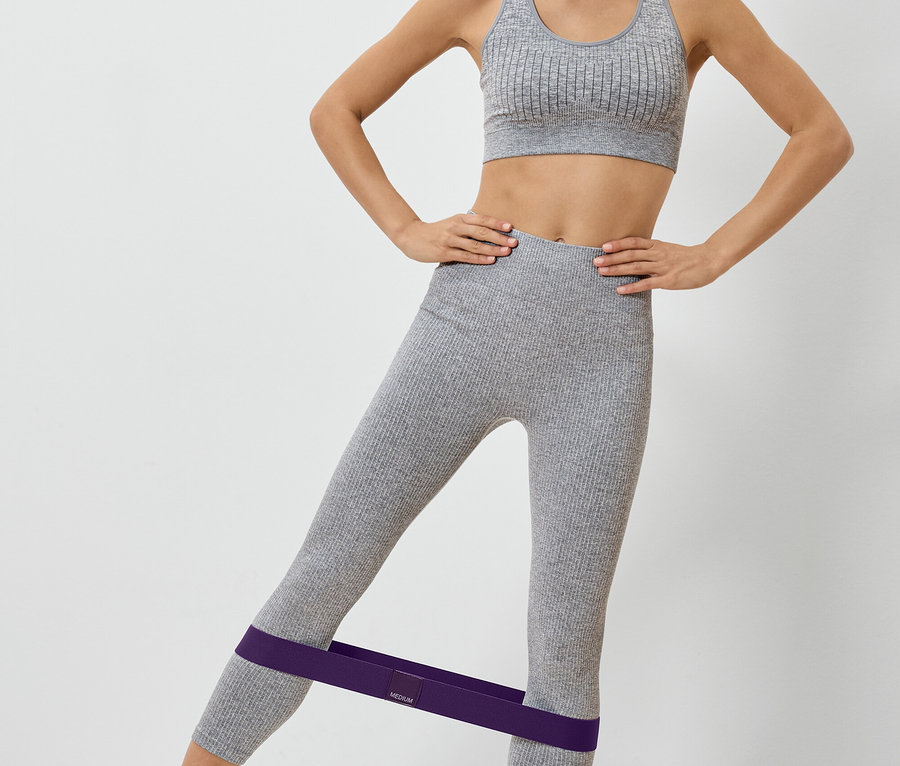 Frau in grauer Sportbekleidung mit lila Fitnessband an den Beinen beim Training.